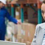 Logistics manager on her laptop in a warehouse, with a male employee checking on boxes in the background.