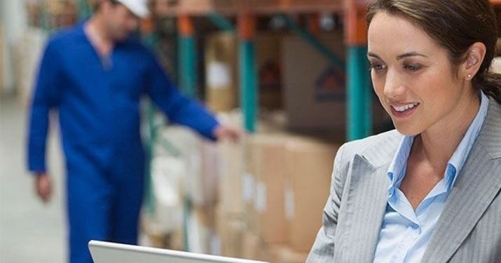 Logistics manager on her laptop in a warehouse, with a male employee checking on boxes in the background.