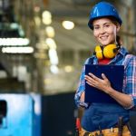 Employee at an industrial facility carrying a clipboard and wearing a blue hard hat and yellow headphones works on optimizing the company's supply chain.