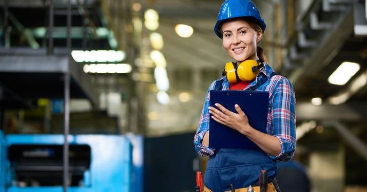 Employee at an industrial facility carrying a clipboard and wearing a blue hard hat and yellow headphones works on optimizing the company's supply chain.