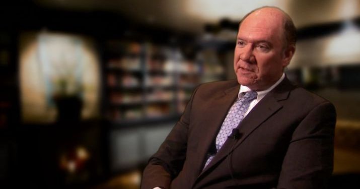 A man in a dark suit and blue checkered tie sitting down talking into a camera about potential reasons for GM's bankruptcy in front of an out of focus library.