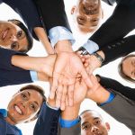 A group of employees in business attire standing in a circle, looking down into the camera and placing their hands on top of each other to show the importance of teamwork.
