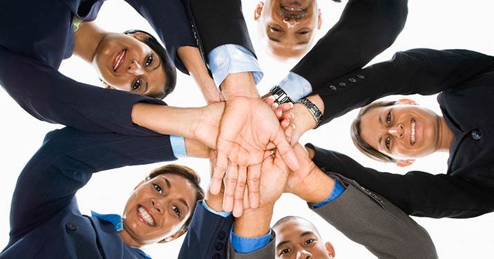 A group of employees in business attire standing in a circle, looking down into the camera and placing their hands on top of each other to show the importance of teamwork.