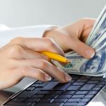Close up of a woman's hands typing on a keyboard searching for high paying jobs while holding a yellow pencil in her right hand and hundred dollar bills in her left.