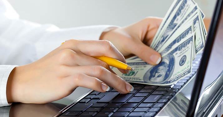 Close up of a woman's hands typing on a keyboard searching for high paying jobs while holding a yellow pencil in her right hand and hundred dollar bills in her left.