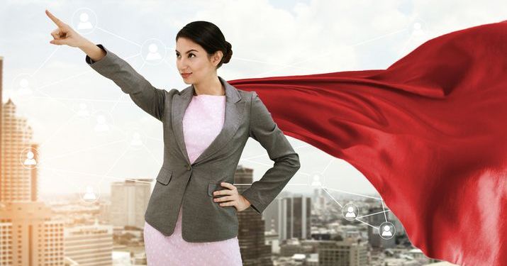 A woman in business attire and a red superhero cape pointing her finger in the air with the backdrop of a city behind her.