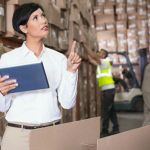 Manager in e-procurement holding a tablet in a warehouse filled with boxes while two warehouse employees work behind her, one in a high-vis vest and the other in a tan uniform.
