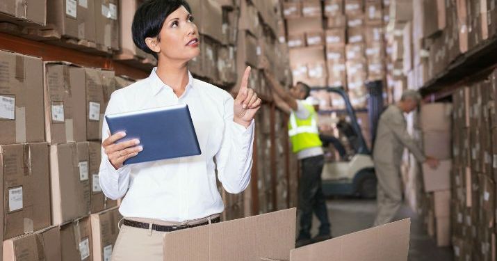 Manager in e-procurement holding a tablet in a warehouse filled with boxes while two warehouse employees work behind her, one in a high-vis vest and the other in a tan uniform.