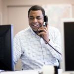 A man smiling and looking at a computer while talking on the phone.
