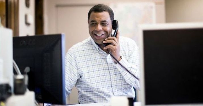 A man smiling and looking at a computer while talking on the phone.