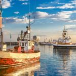 Ships that are part of the seafood supply chain docked in a harbor against the backdrop of a blue sky filled with clouds and a setting sun.