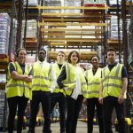A group of workers in yellow reflective work vests in a warehouse with pallets of inventory behind them standing in a row smiling.