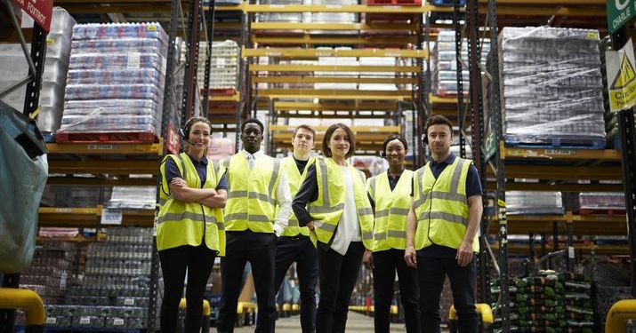 A group of workers in yellow reflective work vests in a warehouse with pallets of inventory behind them standing in a row smiling.