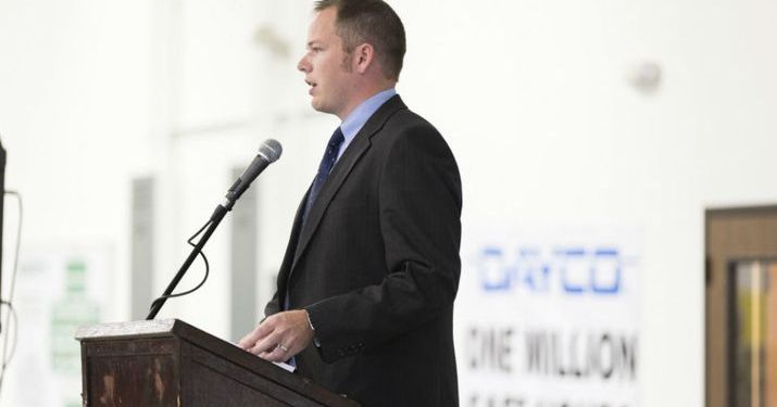 Garrett Stricker speaking at a podium wearing a suit and tie.