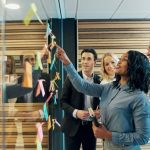 Group of employees working together in an office developing SMART goals by brainstorming and putting post it notes up on a glass.