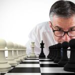 Close up view of a chess board with black and white pieces on each side while a man wearing glasses leans down, staring intently at the chess board trying to understand his competitor's move.