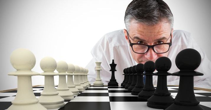 Close up view of a chess board with black and white pieces on each side while a man wearing glasses leans down, staring intently at the chess board trying to understand his competitor's move.