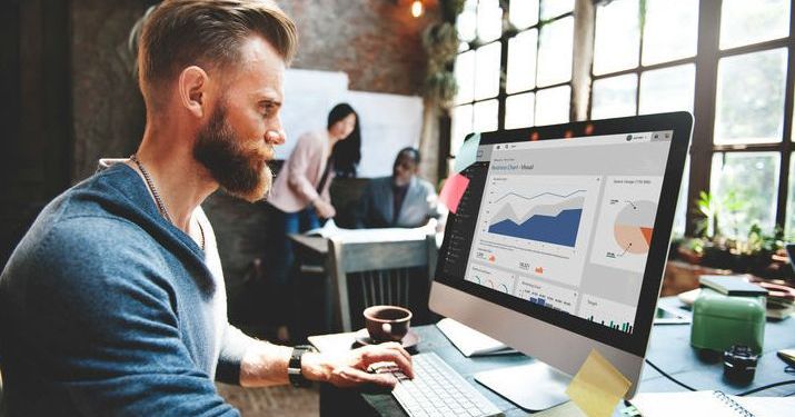 Man with a beard works on large desktop computer looking at different kinds of big data while two other employees are also working in the background.