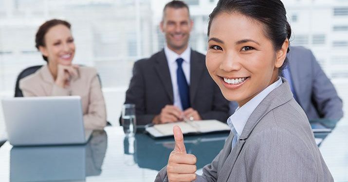 Four employees sitting at a conference room table developing global business strategy, with one woman looking into the camera and giving a thumbs up.