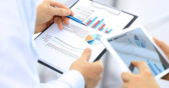 Close up of a pair of hands holding a clipboard with documents showing different types of charts, while another employee is holding a tablet next to him.