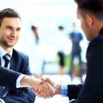 Two men in business suits smiling and shaking hands across a table.