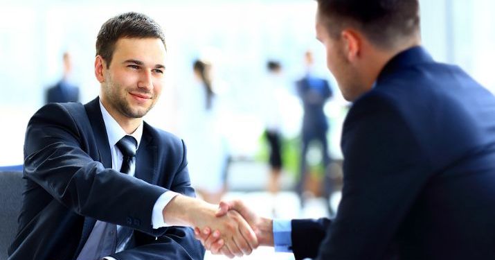 Two men in business suits smiling and shaking hands across a table.