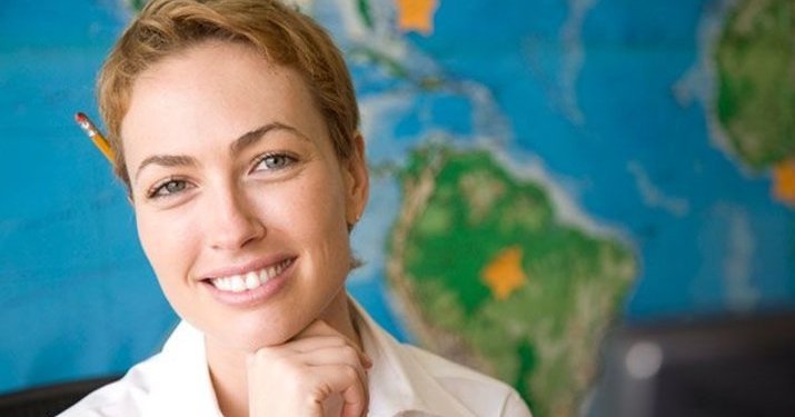 Woman with a yellow pencil behind her ear smiling into the camera in front of a world map.