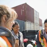 A group of women wearing reflective work vests in a shipyard surrounded by metal shipping containers, working on optimizing their supply chain.