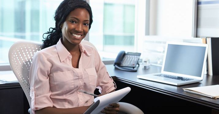 Woman holding a notepad and smiling at her desk in an office, with a laptop on the desk in front of her.