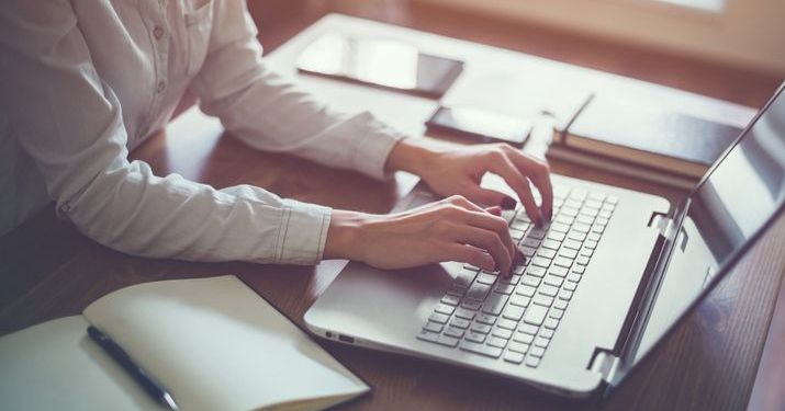 Close up of an online master's degree student's hands typing on a laptop that's sitting on a desk with notebooks, a tablet and a smartphone.