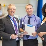 Two healthcare management professionals holding clipboards and smiling, standing with a doctor wearing a stethoscope around his neck.