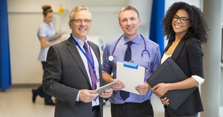 Two healthcare management professionals holding clipboards and smiling, standing with a doctor wearing a stethoscope around his neck.