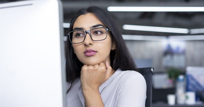 Woman wearing glasses looking a desktop computer screen trying to decide between an MBA and MHA.