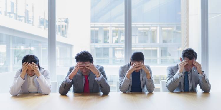Four office employees in business clothes sitting in a row at table with both hands on their heads, feeling stressed out.