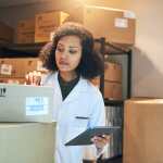 Woman in a white lab coat is checking inventory using a tablet in a storage closet full of cardboard boxes.