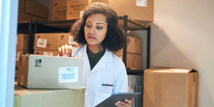 Woman in a white lab coat is checking inventory using a tablet in a storage closet full of cardboard boxes.