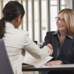 Two women meeting each other and shaking hands across a table.