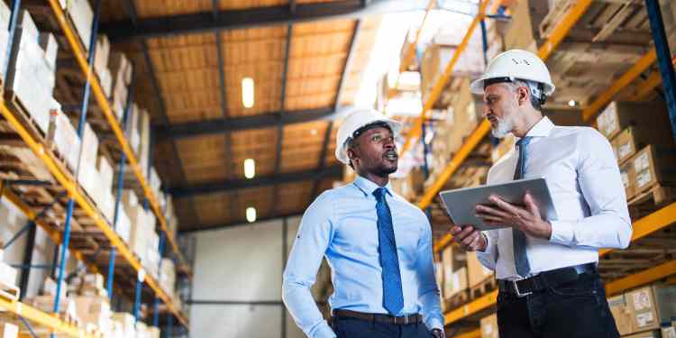 Two men wearing business clothes and white hard hats talking about supply chain in a warehouse filled with boxes of inventory.