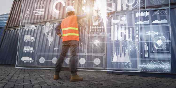 Man in an orange reflective vest and hard hat looking at metal shipping containers that have supply chain graphics and data holographically displayed on them.