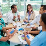 Group of medical professionals sitting around a round table, looking at paper with pie charts and discussing the data.