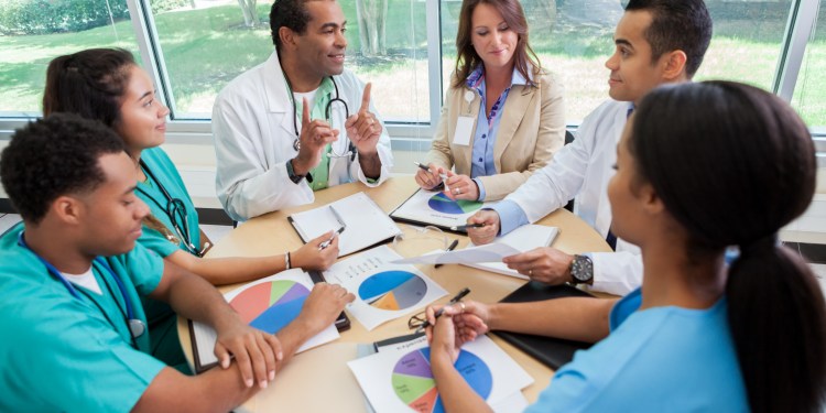 Group of medical professionals sitting around a round table, looking at paper with pie charts and discussing the data.