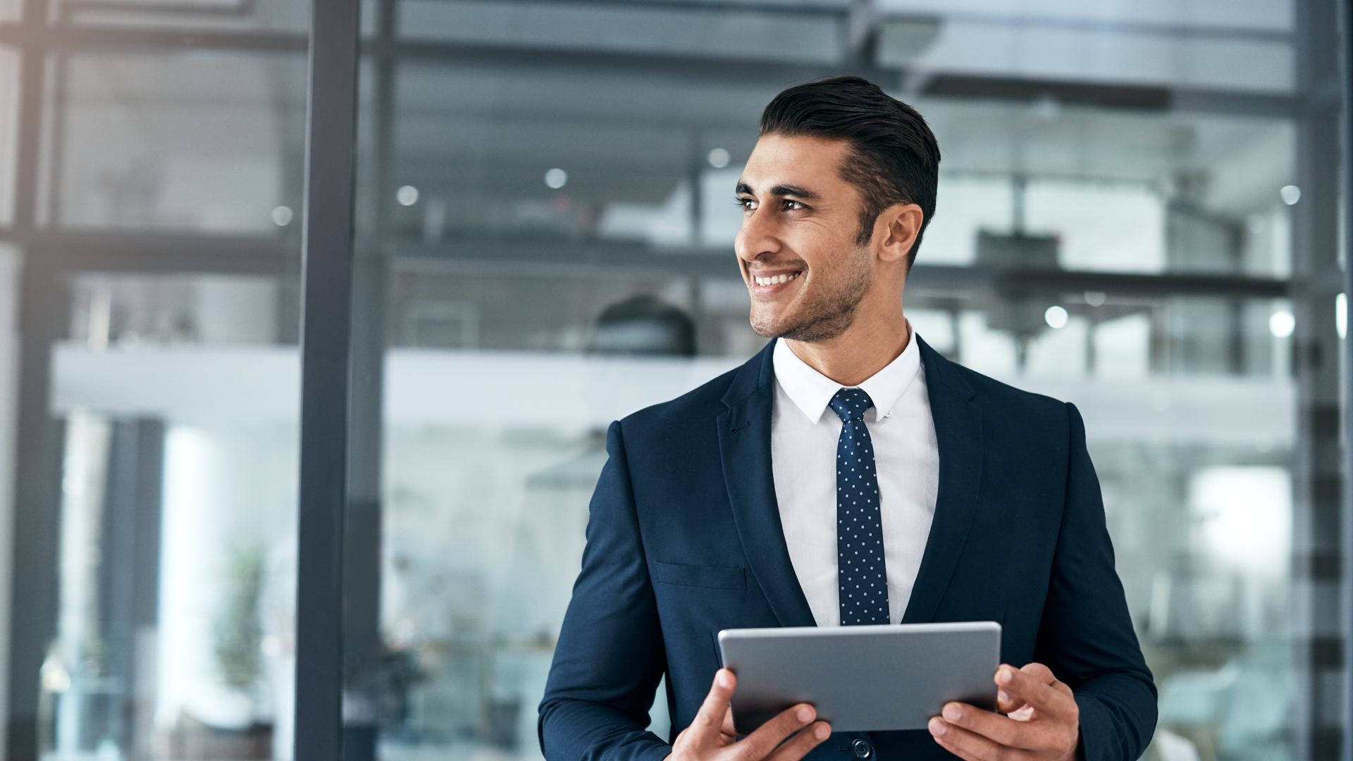 Business man smiling and looking out of an office window while holding a tablet.