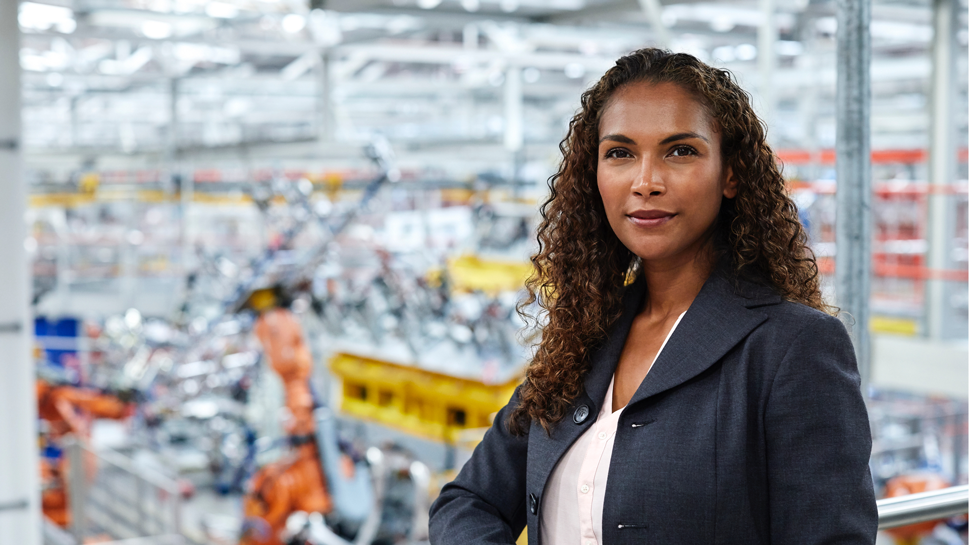 Woman in business clothes standing in a production facility, with heavy machinery out of focus in the background.