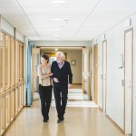 A young woman walking down the hall with an elderly man while holding his arm for support.