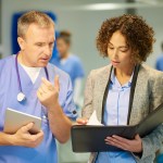 Female healthcare compliance manager holding binder and pointing to papers in binder while having discussion with a doctor in scrubs.