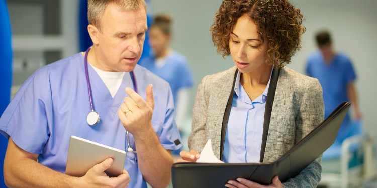 Female healthcare compliance manager holding binder and pointing to papers in binder while having discussion with a doctor in scrubs.