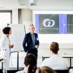 Physician and healthcare administrator presenting concepts via whiteboard and computer projection to medical staff at hospital.
