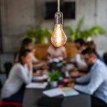 Team of employees at a table working together in the background with a light bulb above in the foreground, symbolizing their innovative teamwork.