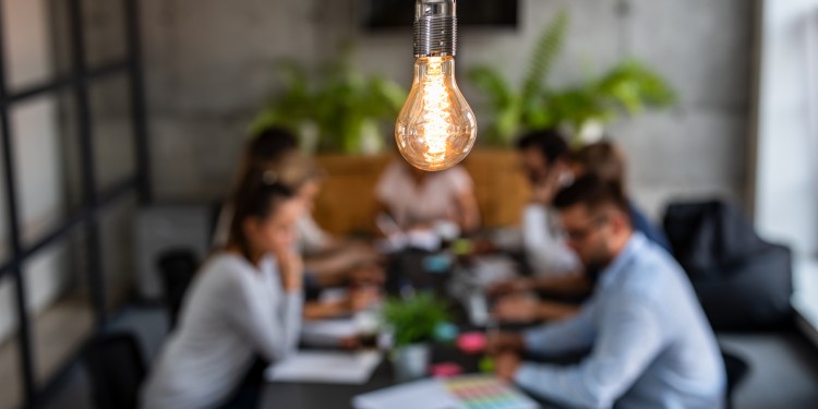 Team of employees at a table working together in the background with a light bulb above in the foreground, symbolizing their innovative teamwork.