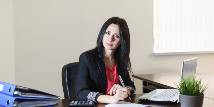 Female administrative services manager in a business suit taking notes on notepad at desk with stack of binders next to her.